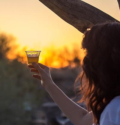 At the treehouse, a young woman holds up a beer sample in a recyclable translucent cup, with a blurred saguaro-studden hillside and sunset in the background.
