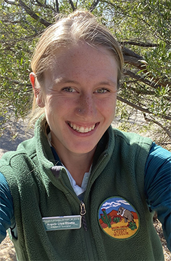Angela stands in a desert landscape, wearing a green fleec vest with an Arizona-Sonora Desert Museum patch on the left side.
