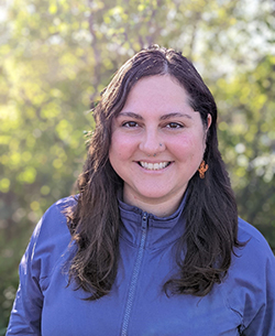 Michelle stands in front of a green-leaved bush, wearing a blue zippered jacket