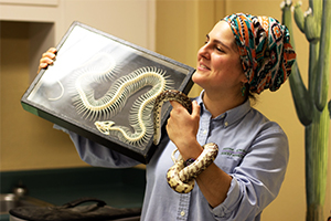 Educator Michelle has a snake wrapped around her left arm and is holding a display case containing a snake skeleton in her right hand