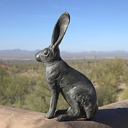 Jackrabbit bronze sculpture sits on an adobe wall with desert landscape and blue sky as background