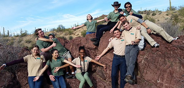 Group of teenagers in a rocky desert landscape, holding their hands up so as to appear to be supporting a large rock above (and behind) them