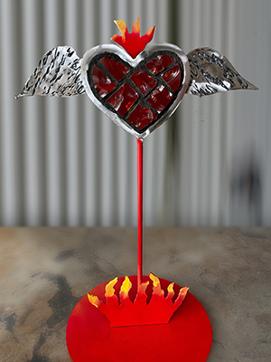 Red Glass Heart with silver wings on the side and and metal flames coming out of the top, resting on a red pole with a base of metal flames